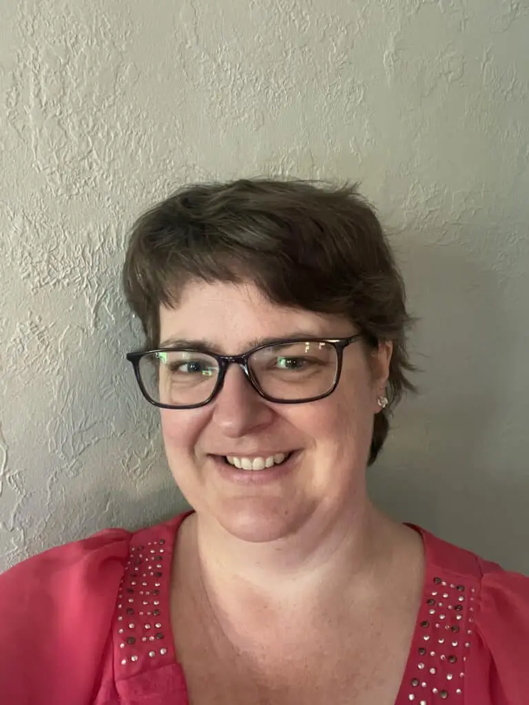 A woman, smiling, with short brown hair, pale skin, purple glasses, and a pink shirt.