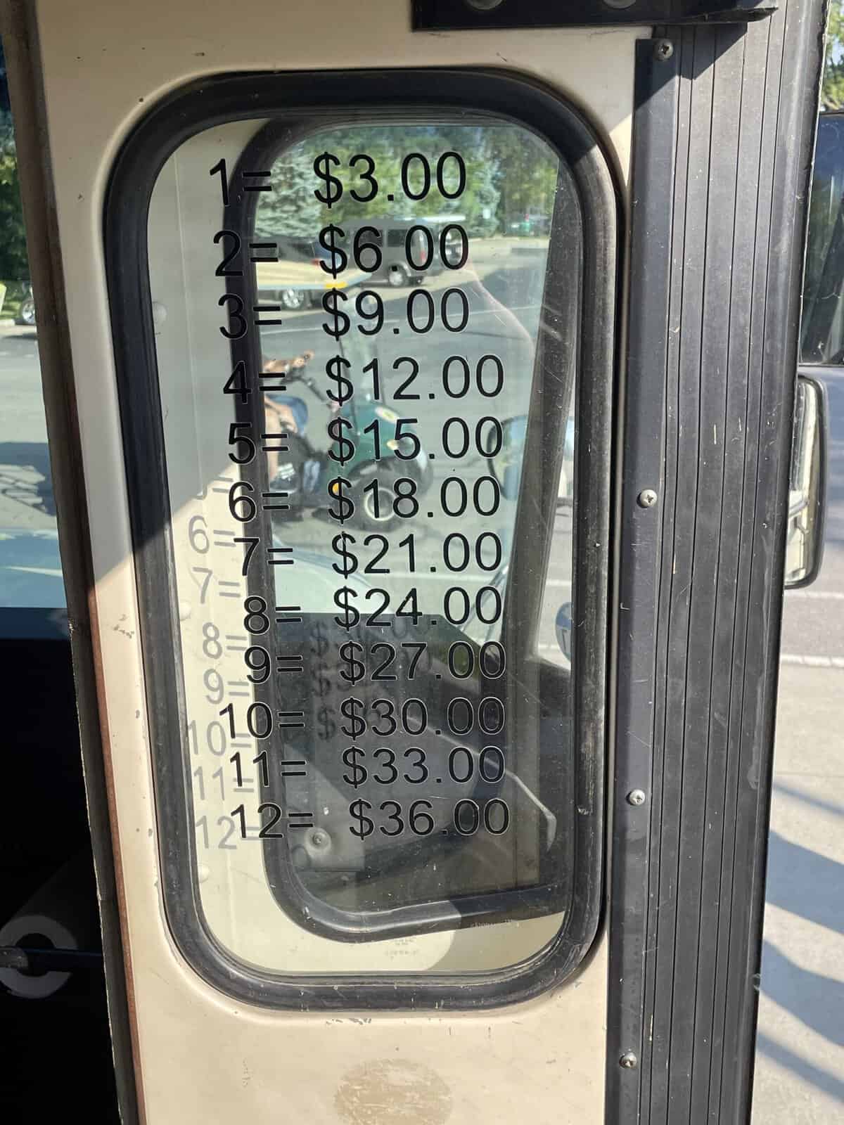 The door of an old school bus converted to a tourist transport in Put-In-Bay, Ohio. On the window of the door is a list of the costs for various numbers of passengers taking a ride. The first line reads 1=$3.00. The second reads 2=$6.00. And so on up through 12=$36.00.